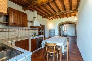 Une cuisine avec des meubles en bois, un mur recouvert de carreaux, une table à manger avec des chaises et une arche en pierre menant à une autre pièce.