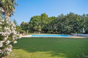 Una piscina rodeada de césped y árboles, con mobiliario exterior y un cielo despejado.