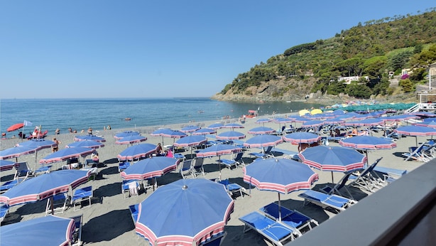 Ein Strand mit vielen Sonnenschirmen und Liegestühlen, ein klarer Himmel und eine hügelige Landschaft im Hintergrund.