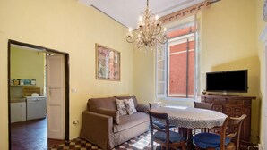 Ein Wohnzimmer mit einer Couch, einem Esstisch mit Stühlen, einer Chandelüster, einem Fernseher und einer Küche im Hintergrund.