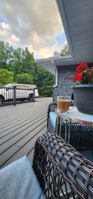 Une terrasse en bois avec des meubles d’extérieur, un véhicule blanc et un pot de fleurs contenant des fleurs rouges.
