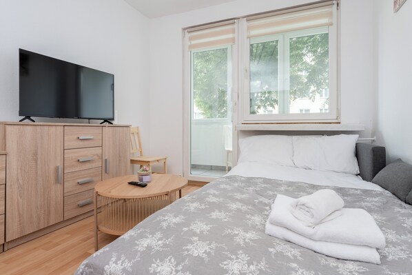 Un dormitorio con una cama, un televisor sobre un mueble de madera, una mesa redonda y una ventana con vistas a árboles.