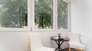 Een kamer met twee witte stoelen, een kleine ronde tafel met een bloempot en grote ramen met uitzicht op bomen.