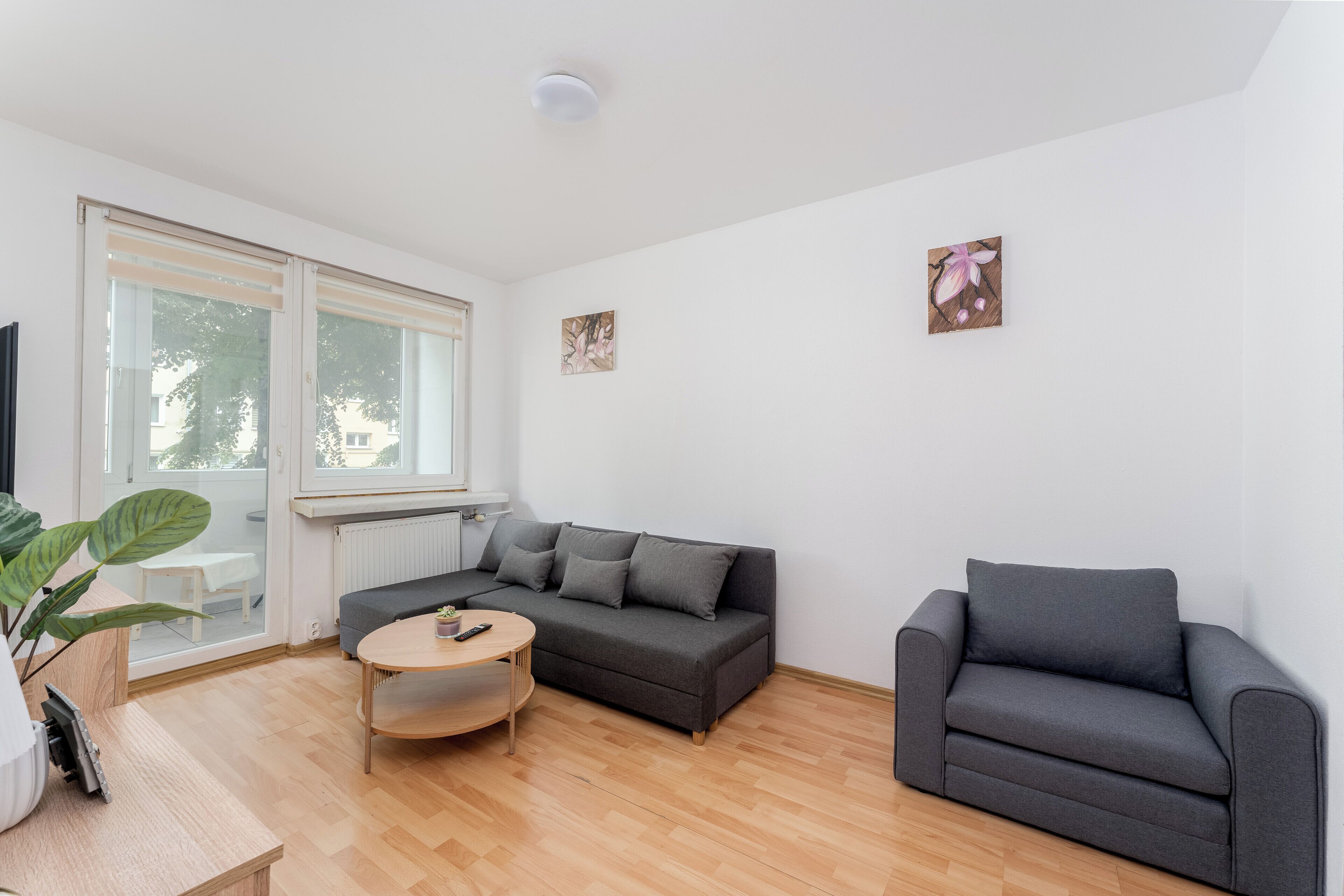 Ein modernes Wohnzimmer mit einem grauen Sofa, einem kleinen Sessel, einem runden Couchtisch und einem großen Fenster.