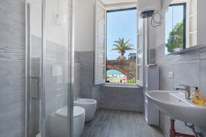 A modern bathroom with a glass shower enclosure, a bidet, a sink, and a window offering a view of a beach and palm tree.