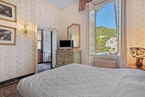 Une chambre à coucher comprenant un lit, une commode, un téléviseur, un miroir et une fenêtre donnant sur une colline et des bâtiments.