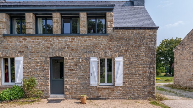 Un bâtiment en pierre avec un toit sombre, une porte et deux fenêtres dotées de volets blancs.
