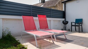 Deux fauteuils de jardin rouge et blanc sont placés sur une terrasse carrelée, avec un barbecue et un fauteuil bleu en arrière-plan.