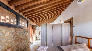 Une chambre avec un mur en pierre, des poutres apparentes en bois au plafond, un lit avec une literie à rayures et une grande fenêtre.