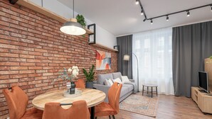 Una sala moderna con pared de ladrillo, sofá gris, sillas naranjas, mesa de comedor de madera y televisor sobre un mueble de madera.