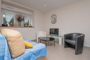 A modern living room with a sofa, a glass coffee table, a black leather armchair, a white chair, a television, and a window with curtains. - Cottage 'Le Cocon' with Private Terrace, Shared Garden and Wi-Fi (Crach)