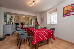 Un comedor con mantel rojo, sillas grises, chimenea de piedra y una ventana con una planta.