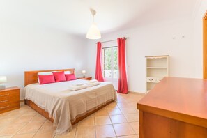 Une chambre à coucher avec un lit, des tables de chevet, un bureau en bois, une fenêtre avec des rideaux et un mur blanc.