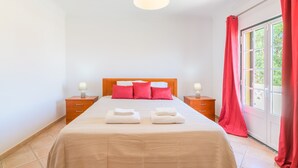 Ein ordentlich bezogenes Bett mit Kissen, ein Nachttisch mit Lampe und ein Fenster mit Vorhängen.