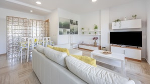 A modern living room with a white sofa, a dining area with a table and chairs, a TV mounted on the wall, and a decorative screen.