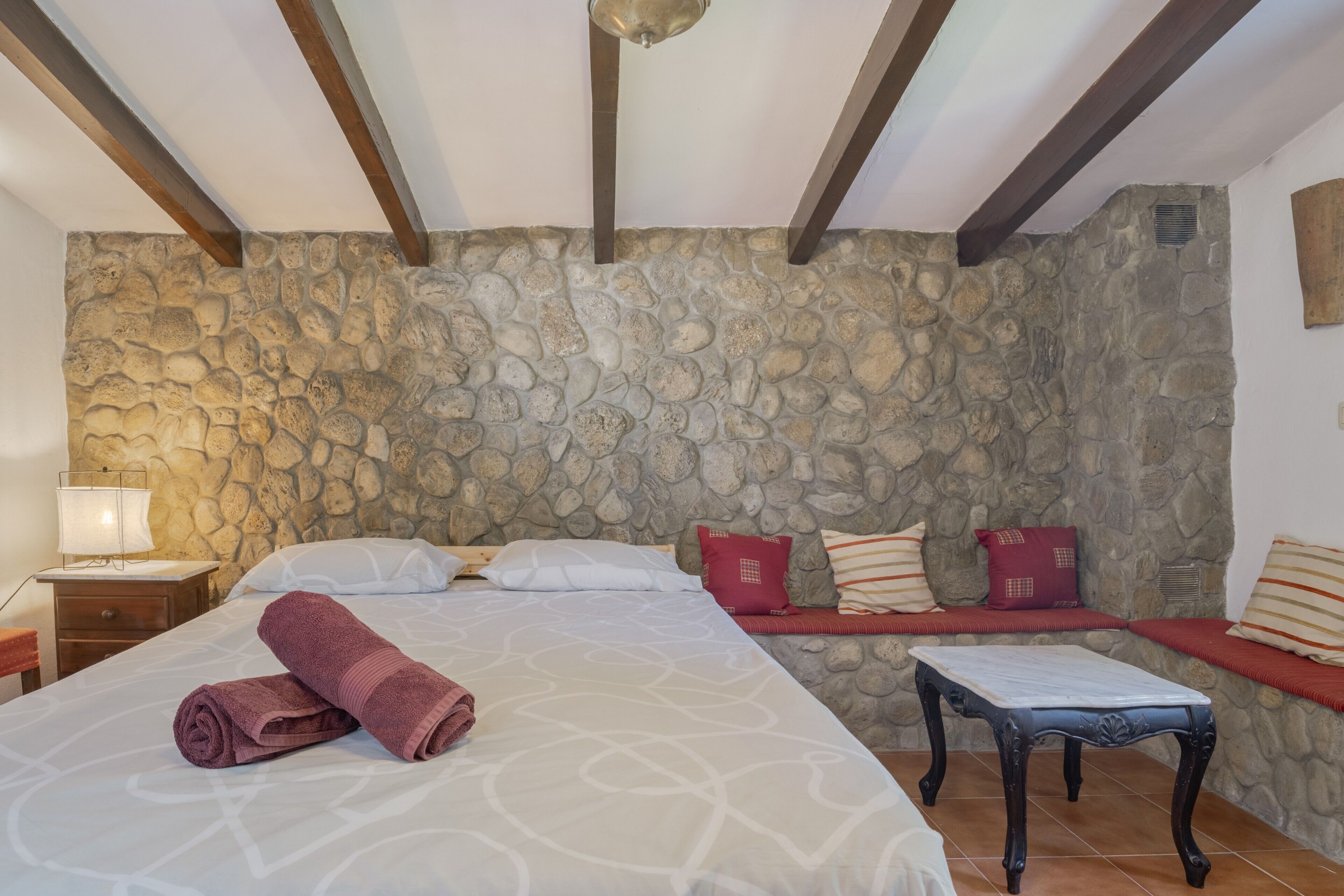 Une pièce avec un mur en pierre, un lit avec une serviette roulée, une table de chevet et un banc avec des coussins.