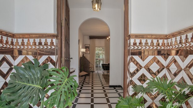 Um corredor com piso xadrez, porta em arco e azulejos decorativos.