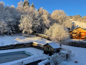 Eine Holzhütte mit Pool, umgeben von schneebedeckten Bäumen und blauem Himmel.