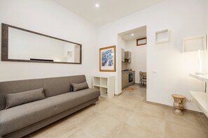 Una sala de estar moderna con un sofá gris, un espejo grande, un cuadro enmarcado y una cocina al fondo.
