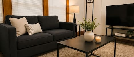 A modern living room with a black sofa, a small table with a vase and candle, a TV, and a window with blinds.