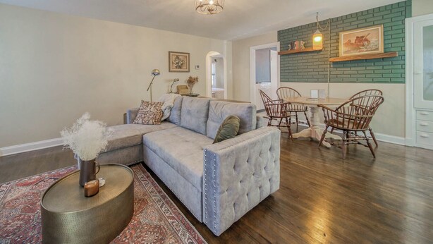 A living room with a sectional sofa, a dining area with wooden chairs, and a kitchen with a brick backsplash. - The Green Acres Cottage - Charming 2 Bedroom near S&T and Acorn Trail in Rolla. (Rolla)