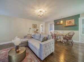 Un salon avec un canapé d’angle, un coin repas avec des chaises en bois et une cuisine avec un mur de crédence en briques.