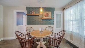 A dining area with a round wooden table surrounded by four chairs, a hanging light fixture, a brick accent wall, and a window with sheer curtains. - The Green Acres Cottage - Charming 2 Bedroom near S&T and Acorn Trail in Rolla. (Rolla)