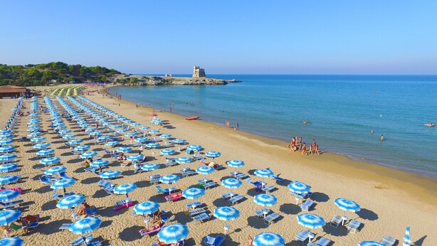 A beach with rows of sun umbrellas and lounge chairs, a clear sky, and a distant view of a castle on a rocky outcrop. - Property In A Holiday Village 'Monolocale 3 Posti Letto' with Shared Pool and Air Conditioning (peschici)