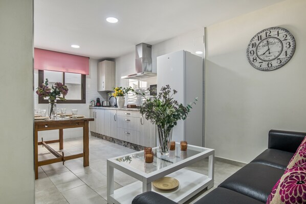 A modern kitchen with white cabinets, a stainless steel range hood, and a dining area with a glass table and a sofa.