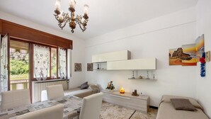 Una sala de estar moderna con comedor, un sofá, una mesa con sillas, una araña, un radiador y un estante de pared con objetos decorativos.