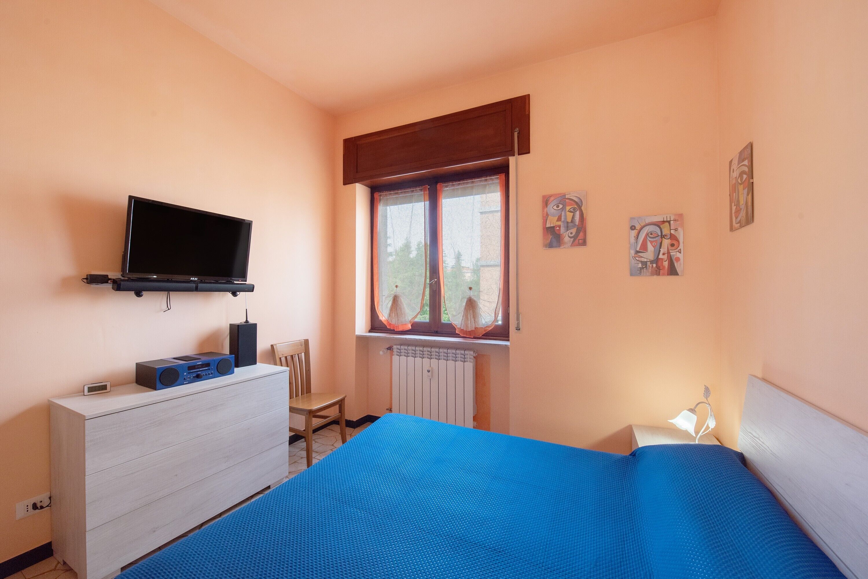 Una camera da letto con un letto, una TV su una cassettiera, una finestra con tende e un quadro appeso al muro.
