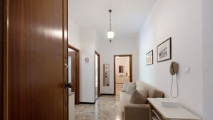 Un pasillo con una puerta de madera, una pared blanca, una lámpara en el techo, un sofá, una mesita y cuadros enmarcados.