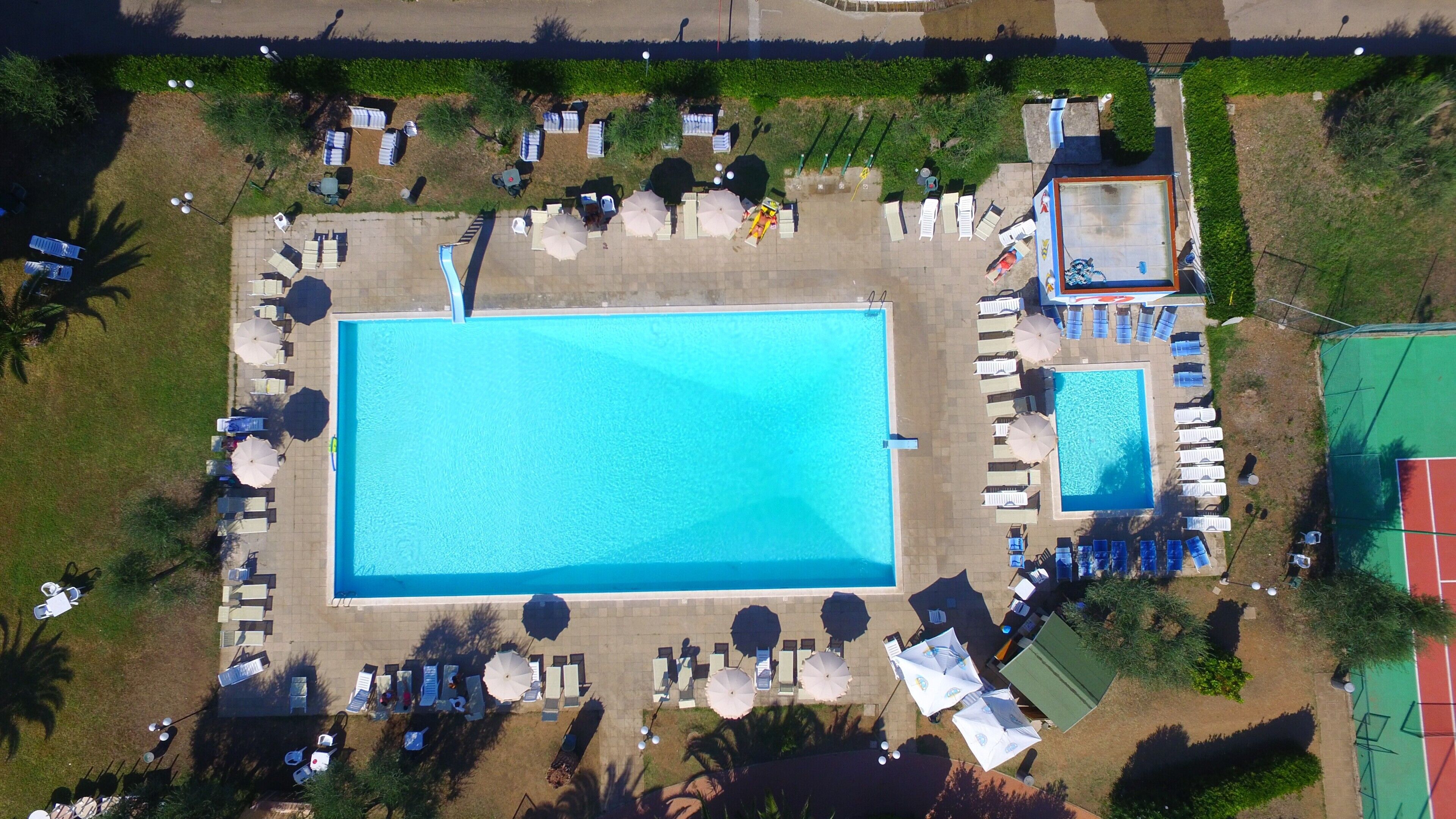 An aerial view of a resort with a large rectangular pool, sun loungers, umbrellas, and a tennis court.