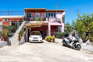 Une maison à deux étages avec une voiture garée dans l’allée, un scooter et un escalier menant à l’entrée.