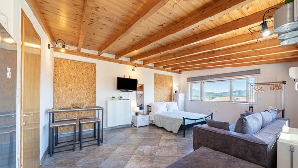 A room with a wooden ceiling, a bed, a sofa, a dining table with benches, a TV, and a window with a view. - Apartment 'Viola' with Mountain View, Wi-Fi and Air Conditioning (Agios Mattheos)