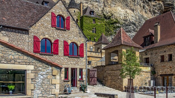 Un edificio de piedra con contraventanas rojas, una escalera y un área de café con terraza.