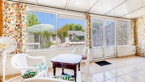 Um pátio ensolarado com mesa e cadeiras, um ventilador e vista para uma casa através de grandes janelas.