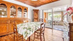 Uma área de refeições com armários de madeira, uma mesa com toalha de mesa florida e vista para um pátio externo através de portas de vidro.