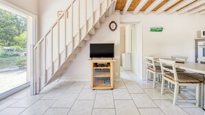 A staircase leading to a second floor, a flat-screen TV on a wooden stand, a dining table with chairs, and a view of greenery outside.