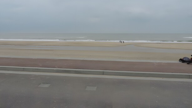 A beach with a road in the foreground and a few people walking along the shore.