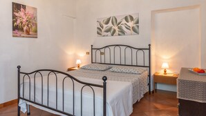 A bedroom with a metal bed frame, a bedside table with a lamp, a small table with fruit, and two wall-mounted paintings.