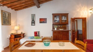 Quarto com mesa de jantar de madeira, cadeiras, cama com telefone vermelho e quadro da Torre Eiffel, armário com louças e um abajur.