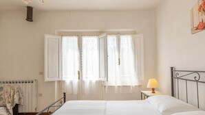 Quarto com cama, mesa de cabeceira com abajur, radiador e janela com cortinas abertas.