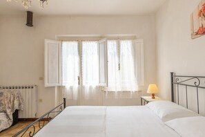 A bedroom with a bed, a nightstand with a lamp, a radiator, and a window with open curtains.