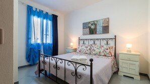 Un dormitorio con cama, cortinas, una mesita de noche, lámpara y un cuadro en la pared.