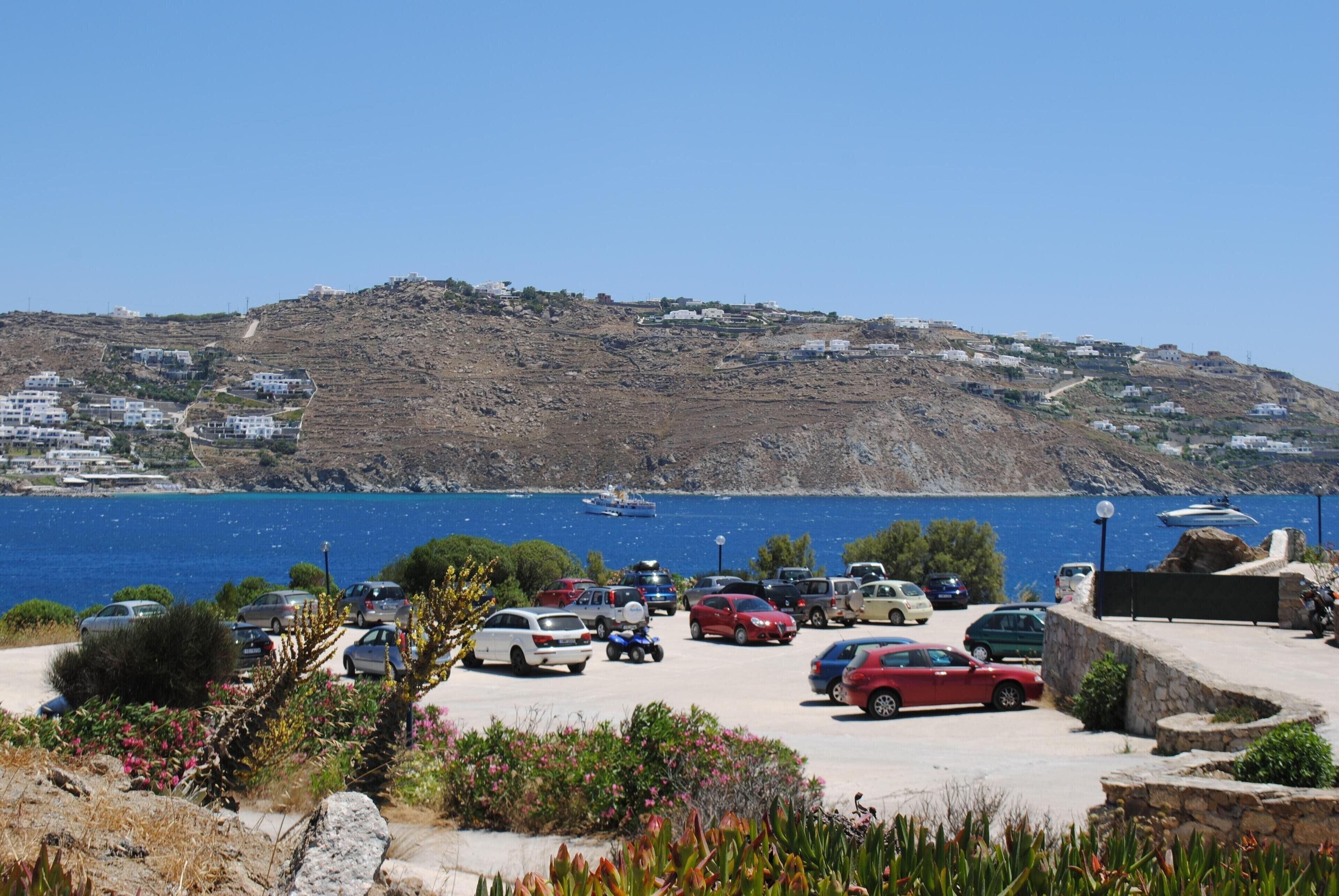 A parking area with cars, a view of the sea, and buildings on a hillside.