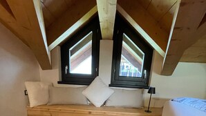 A loft bedroom with a sloped ceiling, wooden beams, a built-in bench with cushions, a fluffy rug, and two windows with shutters. - “Alpine Dream” Apartment in Valmalenco with Mountain Views and Private Garage (Caspoggio)