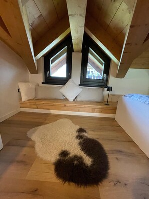 A loft bedroom with a sloped ceiling, wooden beams, a built-in bench with cushions, a fluffy rug, and two windows with shutters. - “Alpine Dream” Apartment in Valmalenco with Mountain Views and Private Garage (Caspoggio)