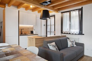 A modern kitchen with wooden beams, a grey sofa with decorative pillows, and a dining table with a patterned tablecloth. - “Alpine Dream” Apartment in Valmalenco with Mountain Views and Private Garage (Caspoggio)