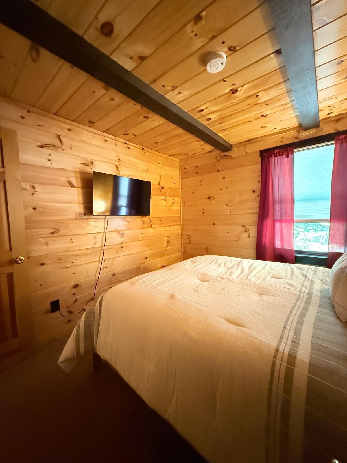 A wooden cabin bedroom with a bed, a mounted TV, and a window with curtains.